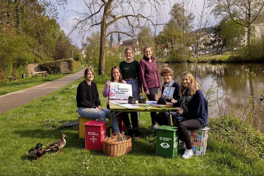 Sechs Frauen sitzen an der Altmühl in Eichstätt um einen Tisch. Sie lächeln. Eine Frau hält ein Plakat des Projektes "Deutschland besser machen" in der Hand.