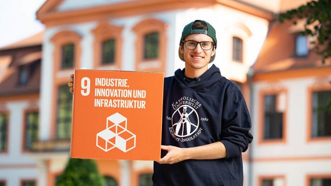 Ein Student mit Brille, Hoodie der Katholischen Universität Eichstätt-Ingolstadt und Cap steht vor der Sommerresidenz in Eichstätt. Er präsentiert einen Würfel mit dem Sustainable Development Goal Nummer 9
