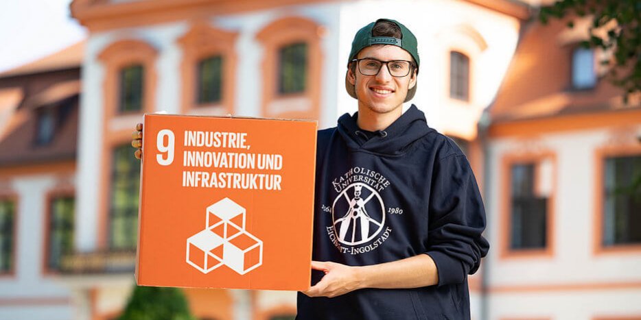 Ein Student mit Brille, Hoodie der Katholischen Universität Eichstätt-Ingolstadt und Cap steht vor der Sommerresidenz in Eichstätt. Er präsentiert einen Würfel mit dem Sustainable Development Goal Nummer 9