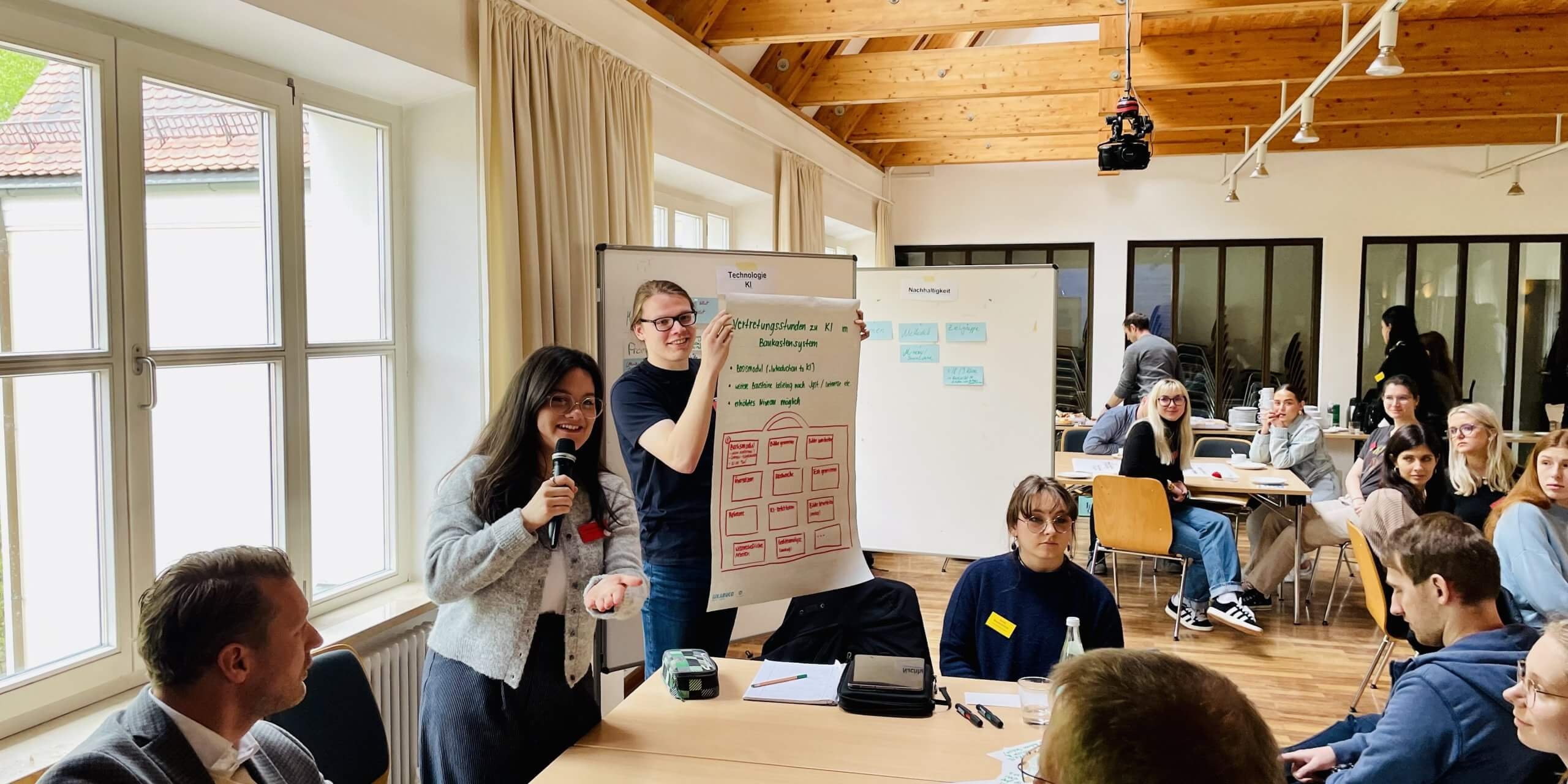 Ein Student präsentiert in einem Seminarraum ein Plakat. Andere Studenten sitzen um einen Tisch und blicken zum Plakat.