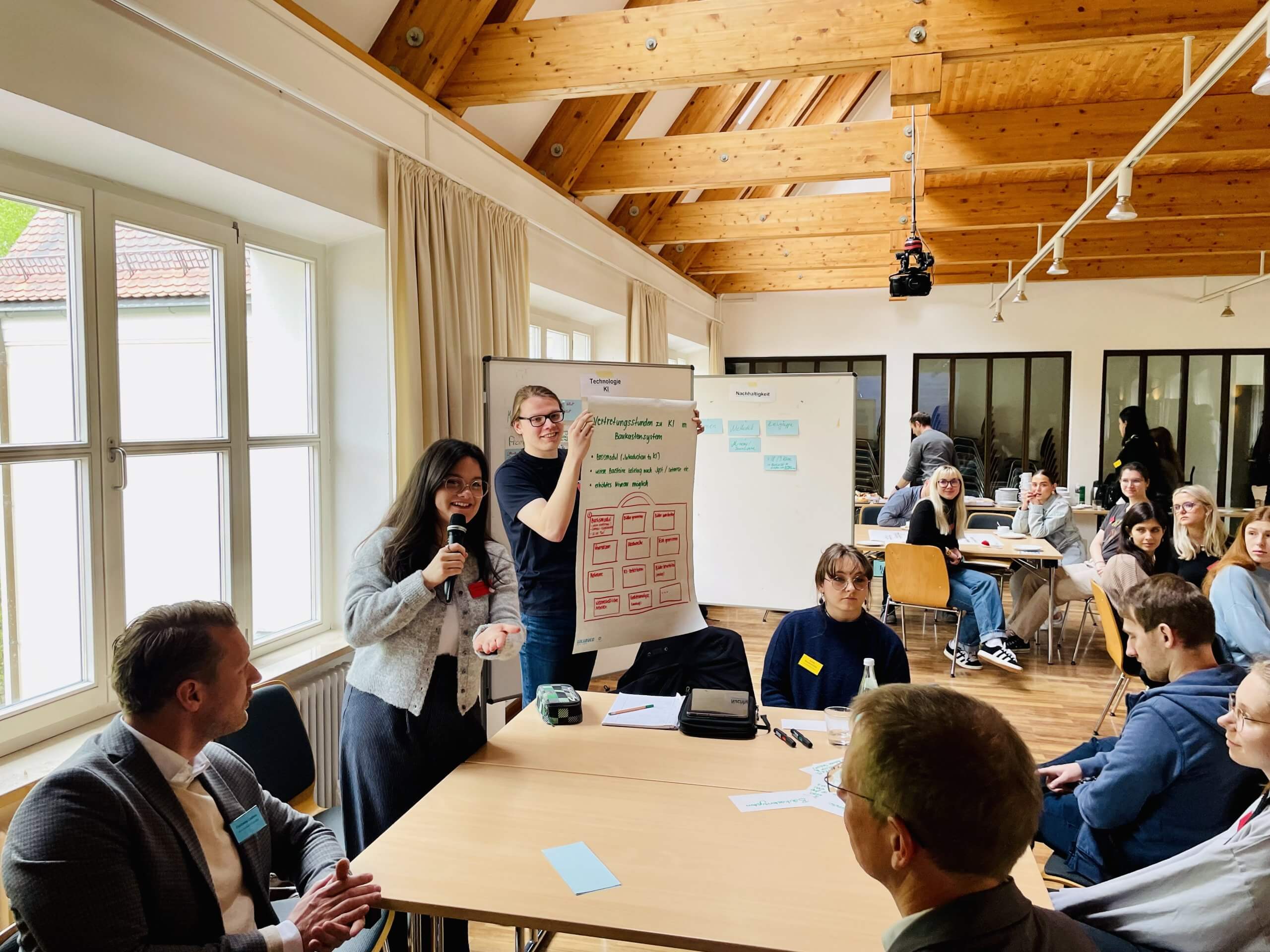Ein Student präsentiert in einem Seminarraum ein Plakat. Andere Studenten sitzen um einen Tisch und blicken zum Plakat.