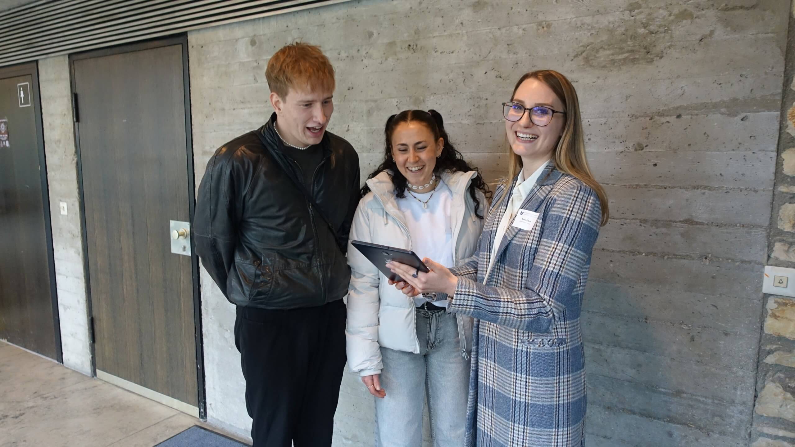 Eine Mitarbeiterin der KU zeigt zwei Studierenden auf einem Tablet das Zukunftsbarometer.