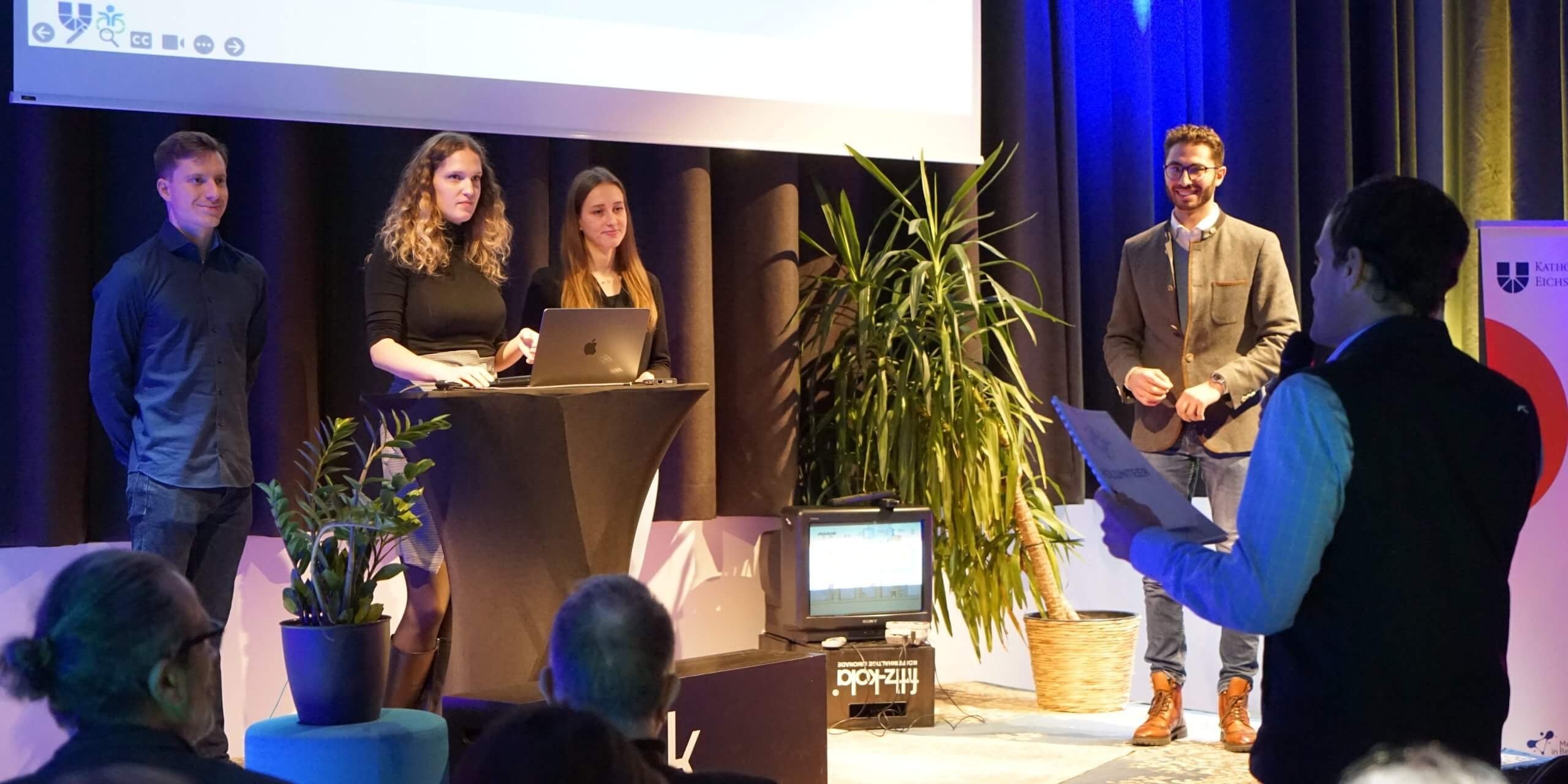 Abschlusspräsentationen WFI Consulting Badge Drei Studierende der WFI stehen mit Johannes Kellner auf einer Bühne. Am rechten Bildrand steht ein Mann mit einem Mikrofon, der zu den Studierenden spricht.