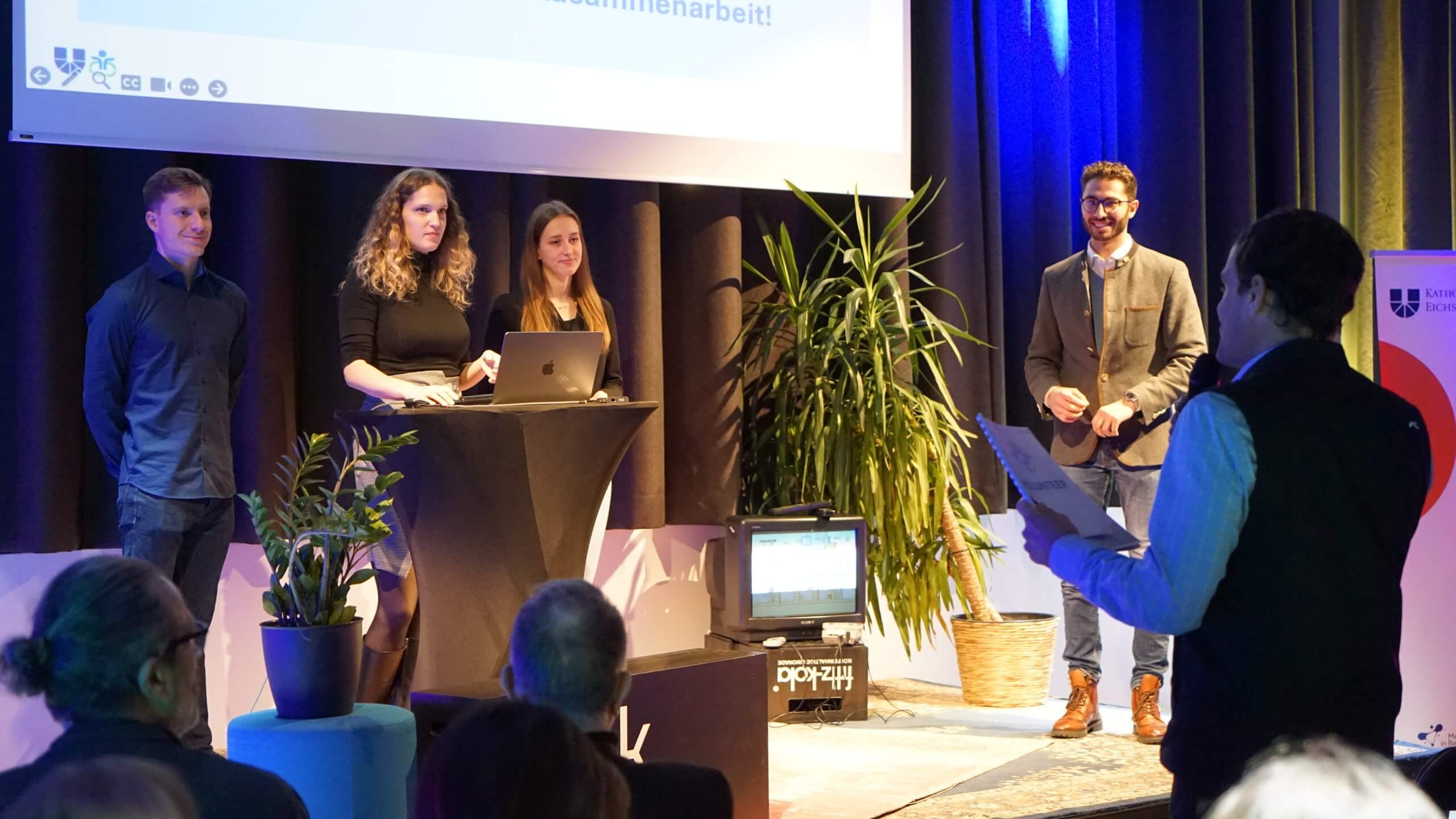 Drei Studierende der WFI stehen mit Johannes Kellner auf einer Bühne. Am rechten Bildrand steht ein Mann mit einem Mikrofon, der zu den Studierenden spricht.